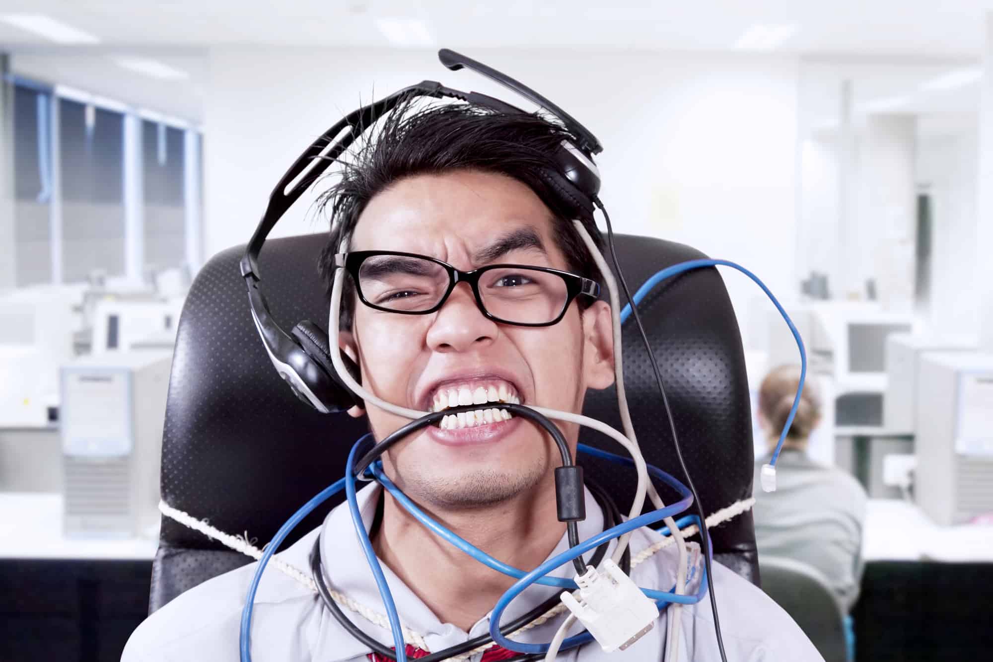 A young Asian man wearing glasses sitting down with headphones on his head. He also is tangled in a bunch of wires, and is biting down of them to express frustration.