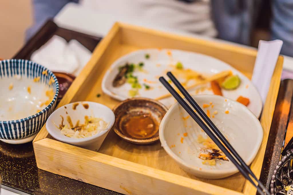 A wooden Japanese lunch box with 4 smaller dishes inside of it with a pair of black chopsticks resting on top of a dish. There is a blue and white bowl on the side of the lunch box. All of the dishes have the remains of food, most of which has been eaten.