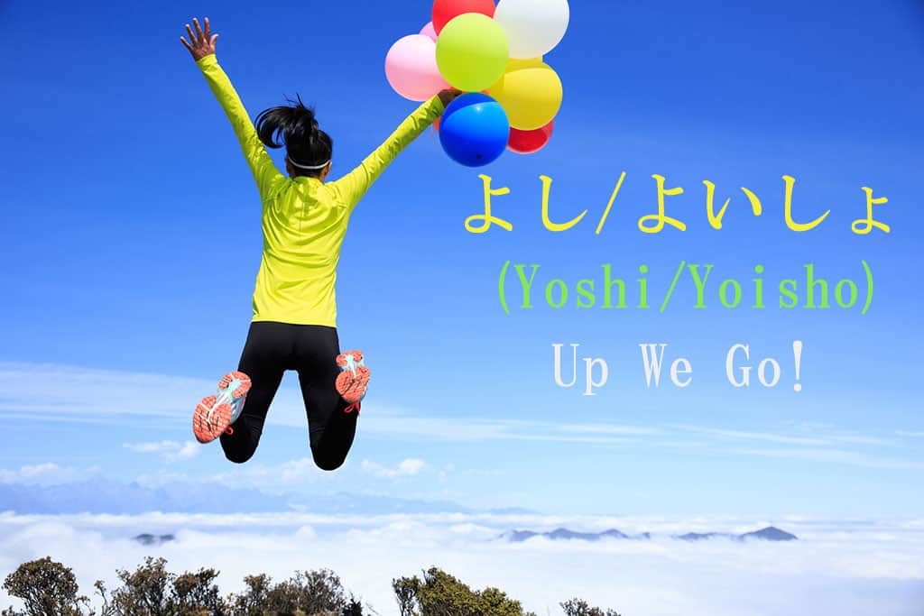 A woman standing at a place of elevation looking over nature (trees and clouds) is holding a bunch of multicolored ballons in her right hand while she is jumping up. The Japanese and English written to the right are for the word "Yooshi-Here-I-go."