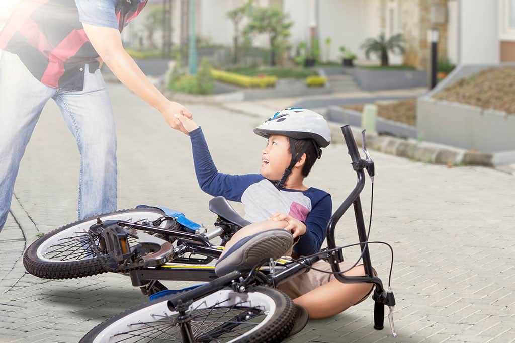 A young boy on the ground after falling down from his bicycle. He is being helped up by someone to the left (a man's hand is seen helping the boy up). The boy is wearing a helmet.
