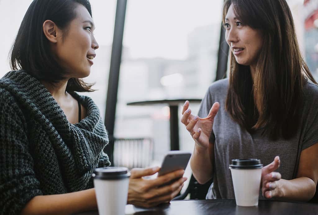 Two young Asian women sitting down at a table with their coffee having a conversation.