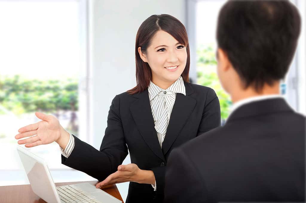 A young Asian woman in business attire using her hands to gesture to a laptop on the table. There is a man in front on her, which we can only see the back of his head.