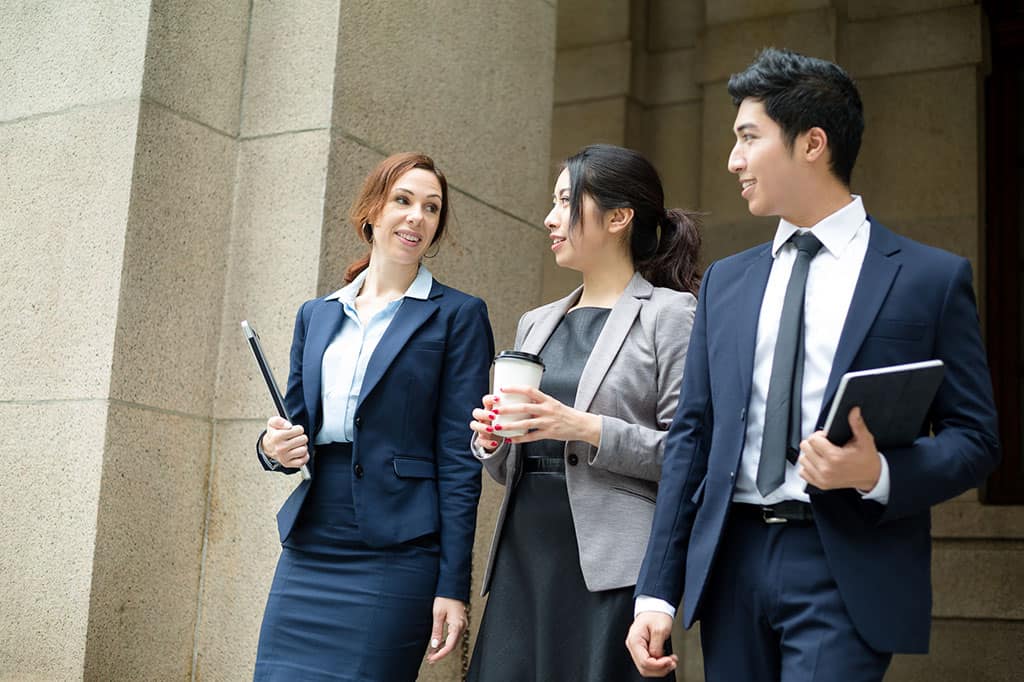 A group of 3 young people in business attire, standing next to each other having a conversation.