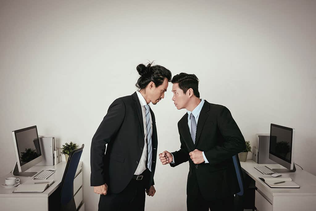 Two Asian men dressed in suits standing face to face with their foreheads pressed against one another. Their desk with a computer on it is behind them, and they look to be angry at each other.