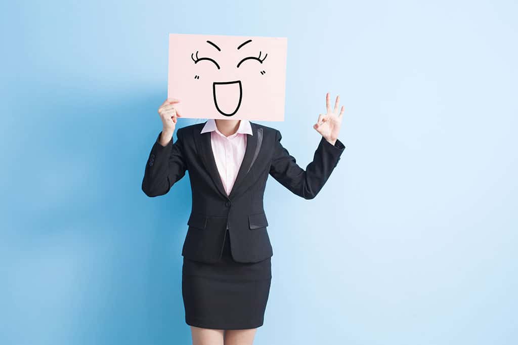 A woman in a business outfit with a pink board that has a happy face drawn on it, which she is holding in front of her face.