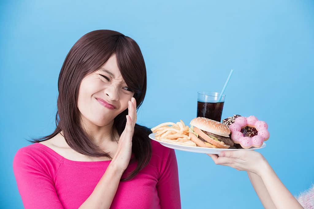 An Asian woman in a pink outfit putting out her hand to refuse a plate full of unhealthy food (french fries, hamburger, donuts, soda).