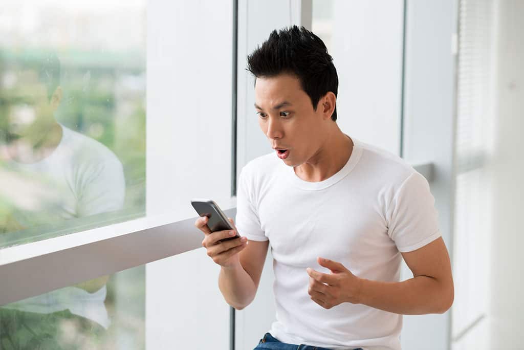 A young Asian man looking at his phone with a surprised look on his face. He is standing next to a window, and a blurred out green background can be seen.