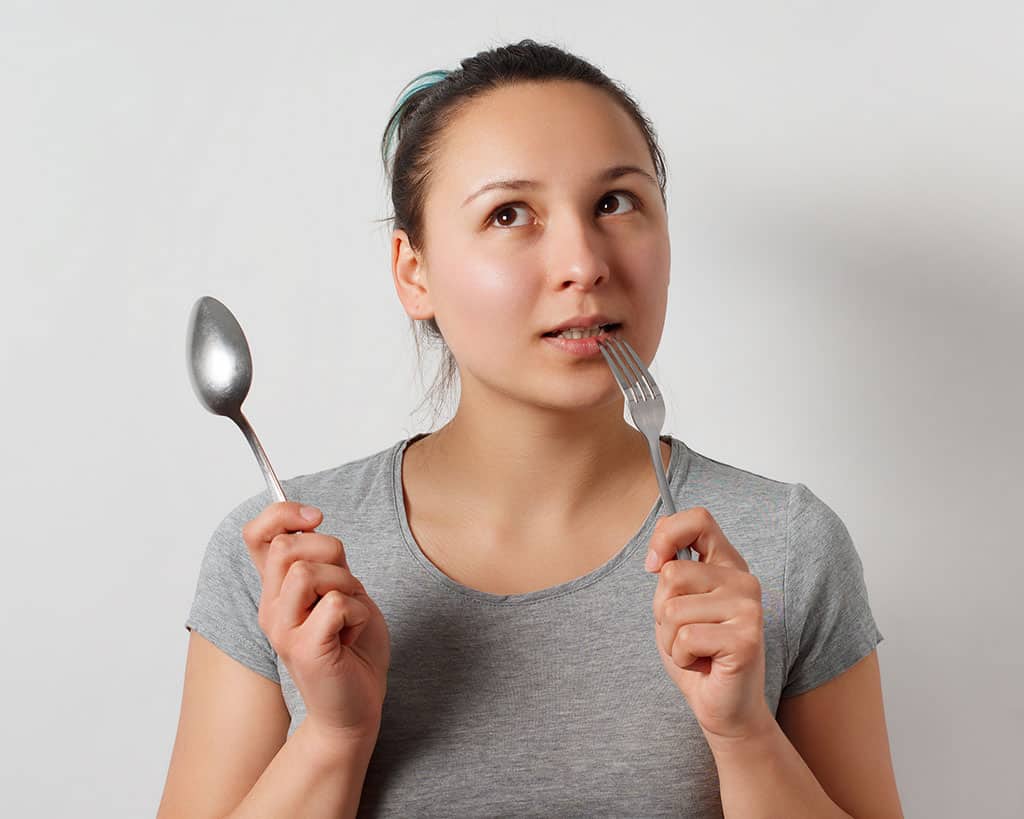 A woman holding a spoon in her right hand, and a fork in her left hand. The fork is in her mouth, and the woman looks like she is thinking of something to eat.