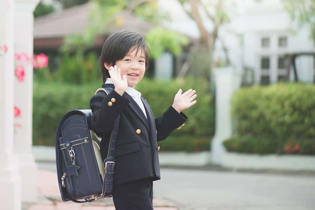 A young, Asian boy dressed in a Japanese school uniform and backpack, waving goodbye.