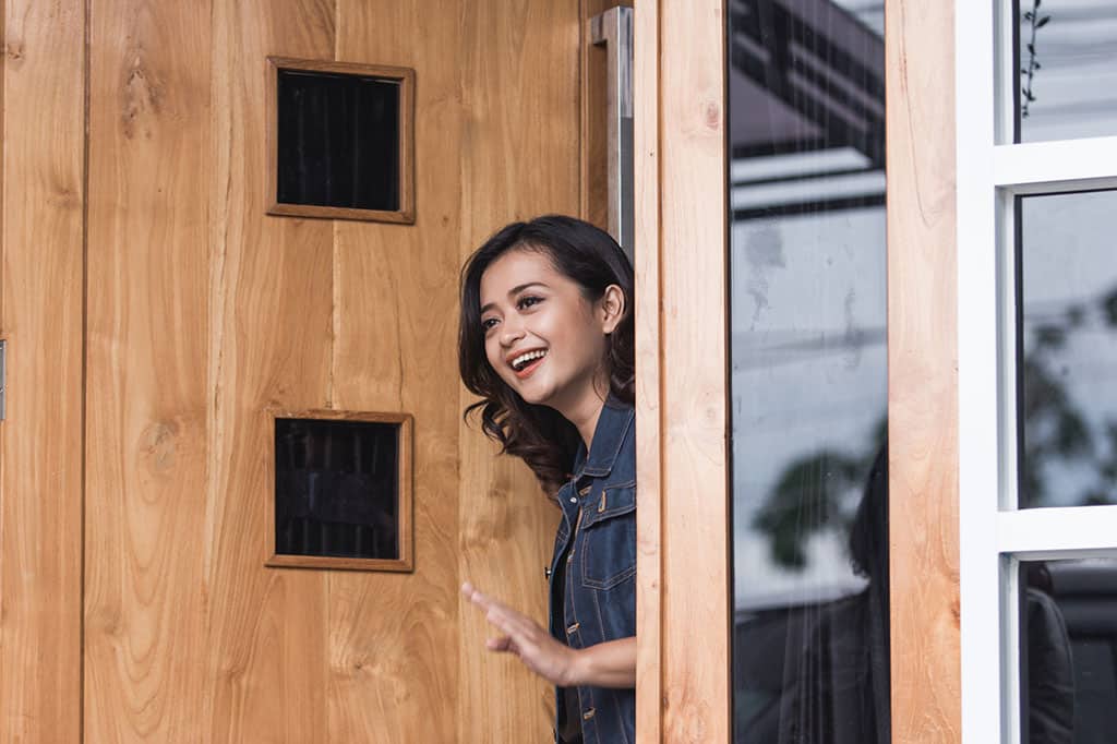 An Asian woman looking out of a door of a home, waving goodbye to someone.