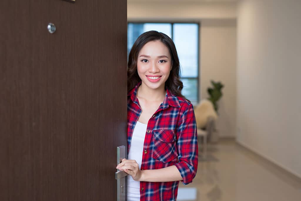 A young, Asian woman standing behind an open door, smiling as if to welcome to someone.