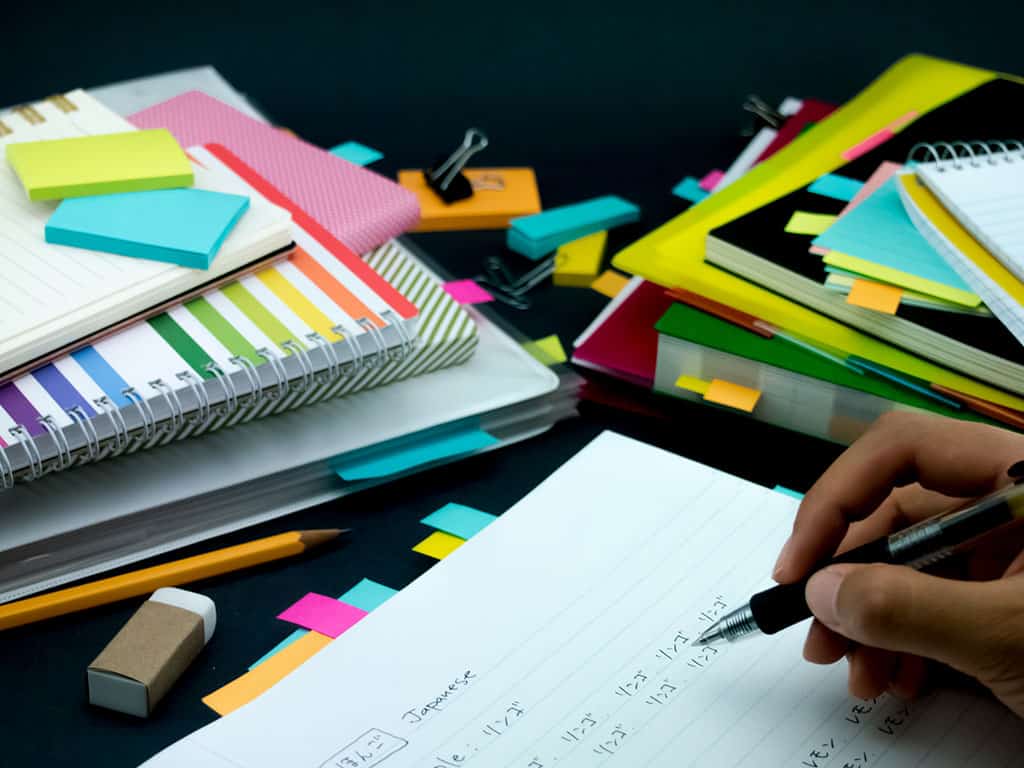A stack of note books, color sticky note tabs, and an open notebook with Japanese written on it. On this open notebook, we can see a hand holding a pen, as if in the process of writing more.