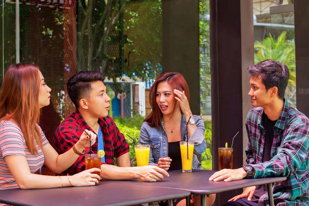 A group of young Asian men  (2) and woman (2) sitting down at a table drinking iced tea and juice, having a conversation.