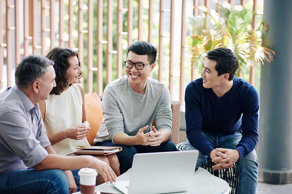 A group of 4 people sitting down around a small table with a laptop on it. There are two young Asian men on the right, and a older white woman and man on the left. They look to be having an enjoyable conversation.
