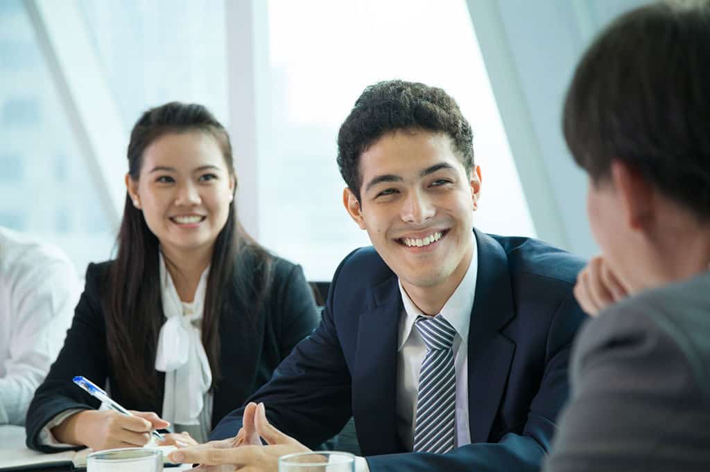 A young man and woman in business attire, sitting at a table speaking to someone off to the right (only the back of his head is visible).