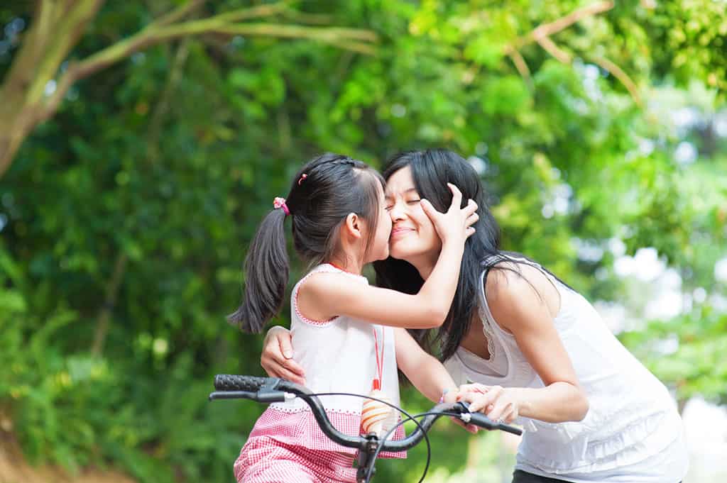 A young girl on a bicycle being held by her mom. She is kissing her mom on the cheek.