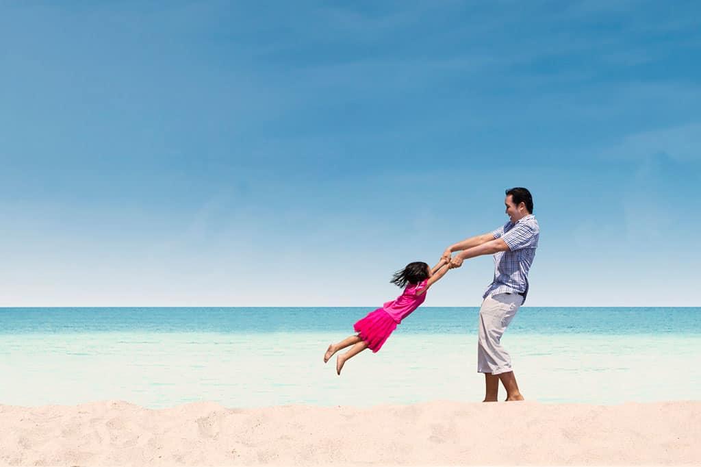 A young man swinging a young girl by her arms on the beach.