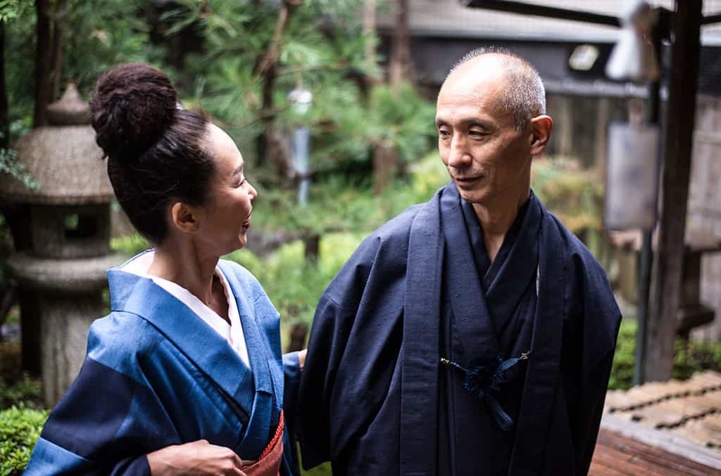 An older woman and man dressed in traditional Japanese clothes, outside in a garden looking at one another.