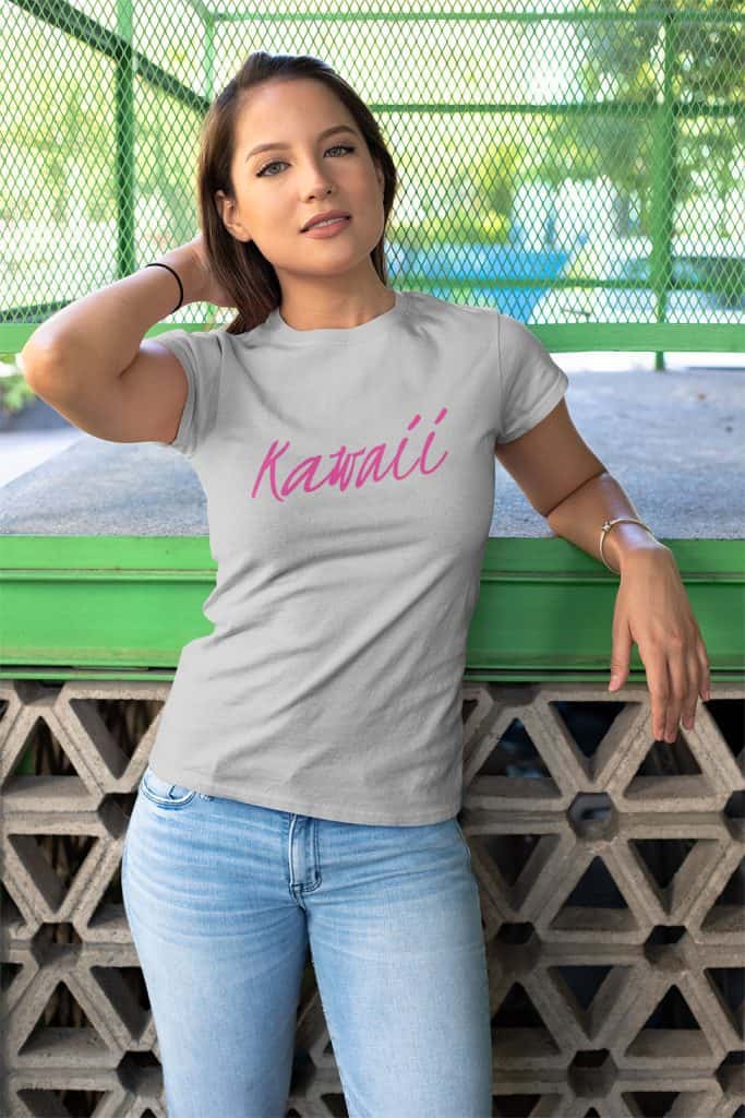 A young woman learning against a ledge. She is a wearing a grey shirt with pink text that reads, "Kawaii" on it.