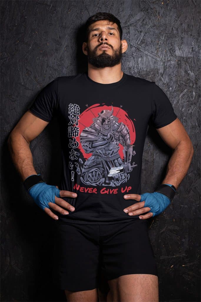 A young man with a beard and blue hand wraps in leaning against a wall wearing a black shirt that says, "絶対諦めない!" and "Never give up."