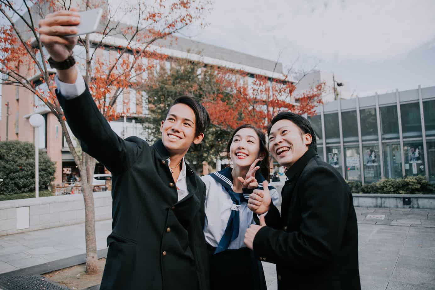 Two young Asian boys, and one young Asian girl, all wearing Japanese high school student uniforms. The boy on the left is holding up a smartphone to take a selfie of them all together.