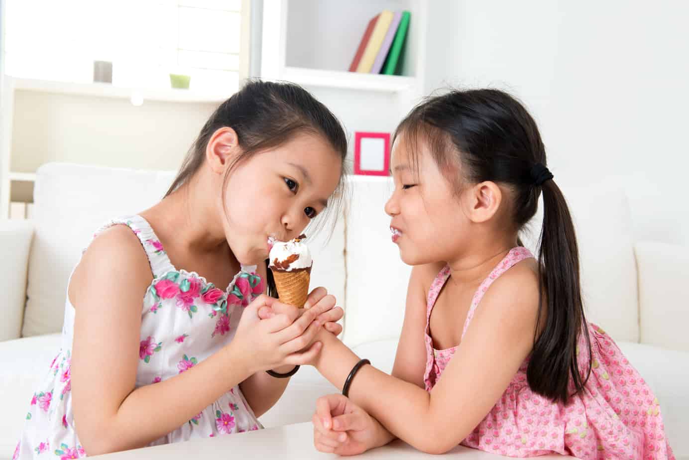 Two young girls in a white room sharing an ice cream cone.