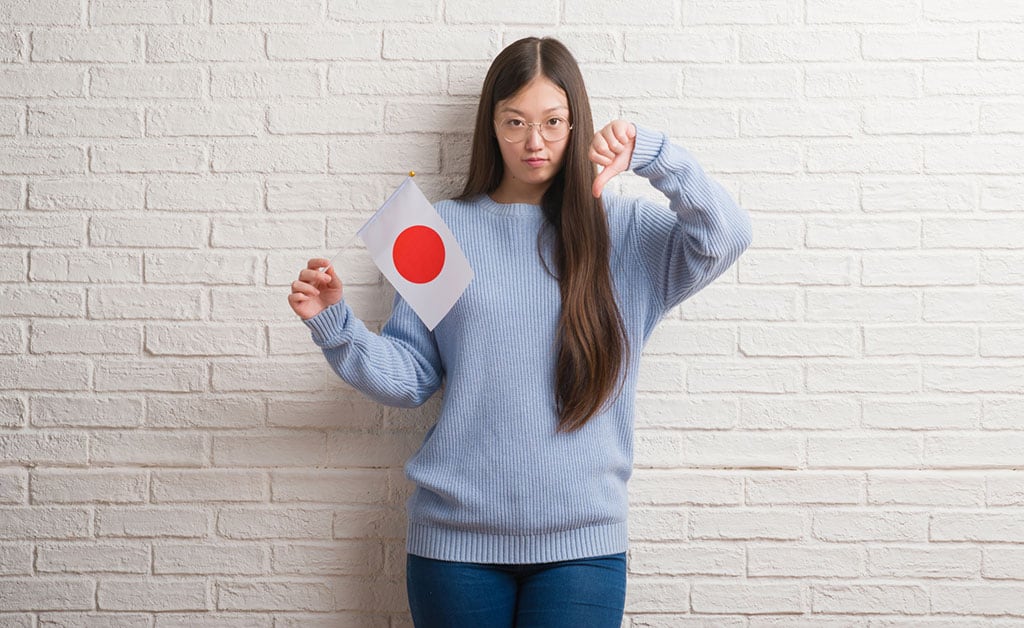 A young, Asian woman holding a small Japanese flag with her right hand and doing the thumbs down gesture with her left hand.