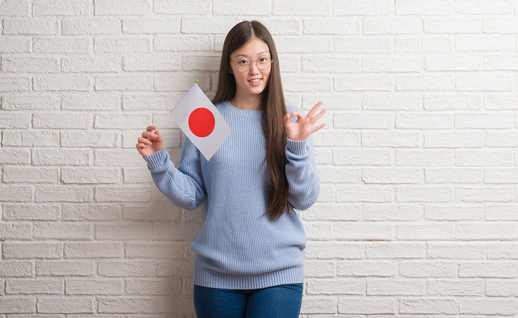 A young, Asian woman holding a small Japanese flag with her right hand and giving the "OK" gesture with her left hand.