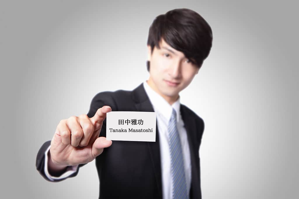 An Asian man in a business suit holding a business card that has Japanese kanji and the English "Tanaka Masatoshi" on it.  