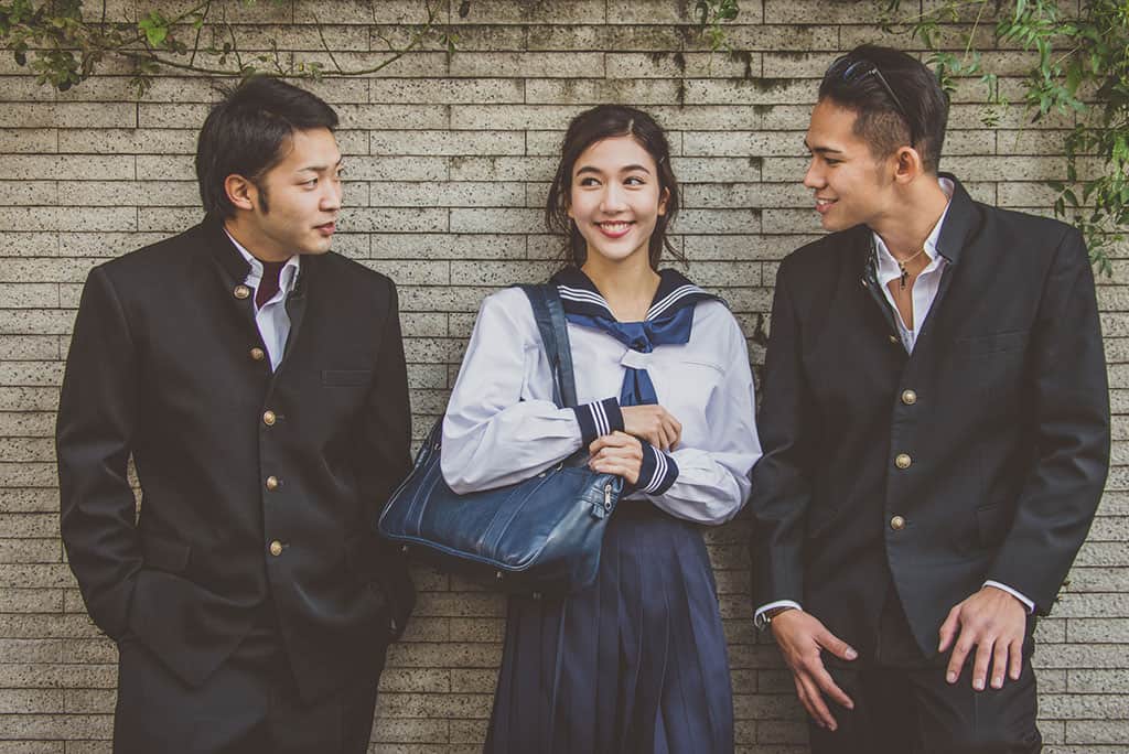 Two Japanese high school boys and one girl all in their school uniforms leaning against a wall talking.