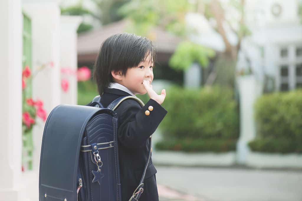 A young Asian boy dressed in a Japanese school uniform and backpack waving to someone.