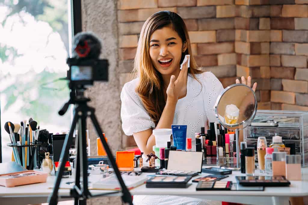 A young, Asian woman is in front of a camera, with make-up products all around her. She is wiping her cheek with a white cloth.