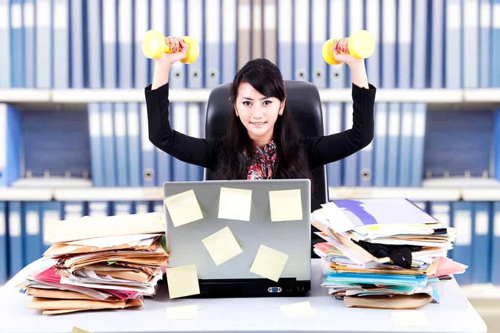 An Asian woman at a messy office desk lifting yellow dumbbells over her head.