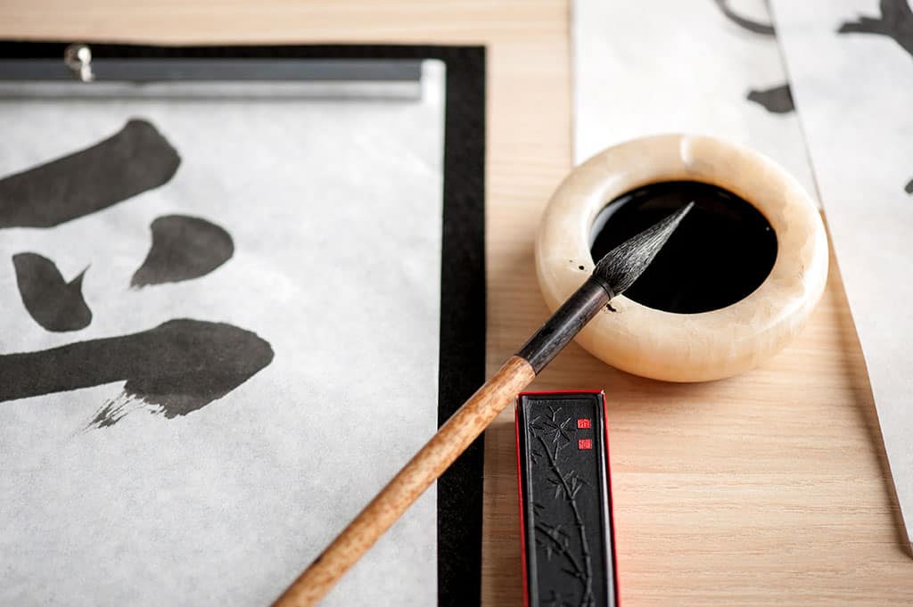 Japanese calligraphy. A brush in resting on a container filled with black ink. Pieces of paper with Japanese characters on it can be seen.