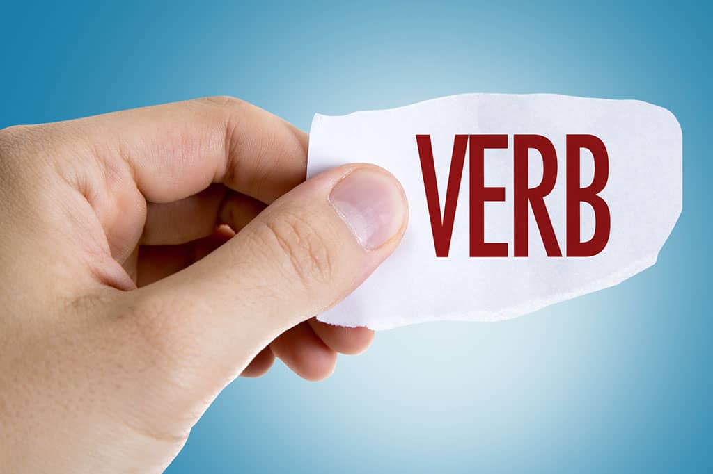 A human hand holding a piece of white paper with the word, "VERB" written on it in red.