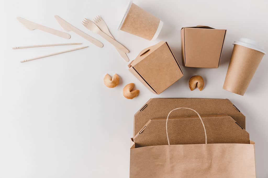A brown paper bag, Chinese take out containers, cups, a pair of chopsticks, knives, forks, and fortune cookies laid out on a table.