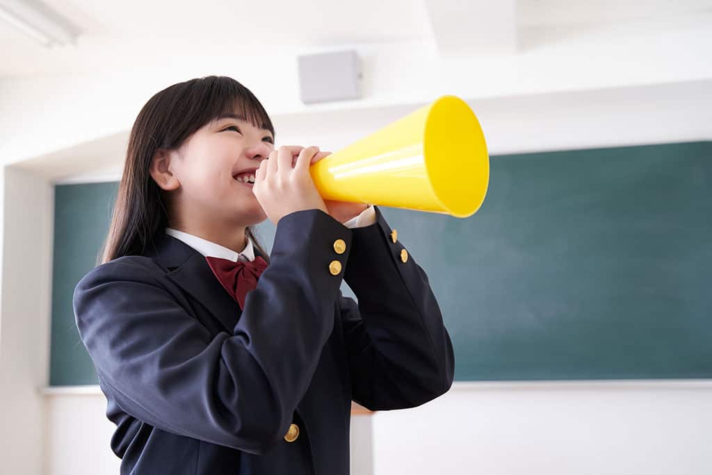 A girl dressed in a Japanese school uniform holding a yellow cheering cone to her mouth looking as if she is cheering.