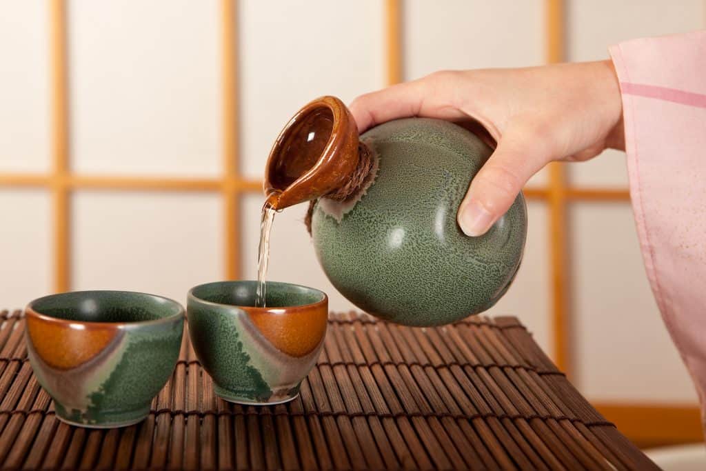A woman's hand is seen pouring sake out of a green Japanese style container into a cup, with another cup next to it.