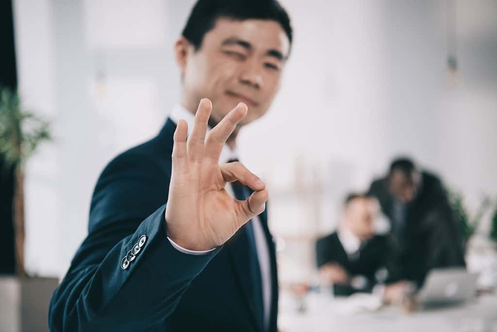 A young, Asian man in a business suit winking and giving an okay gesture with his fingers.