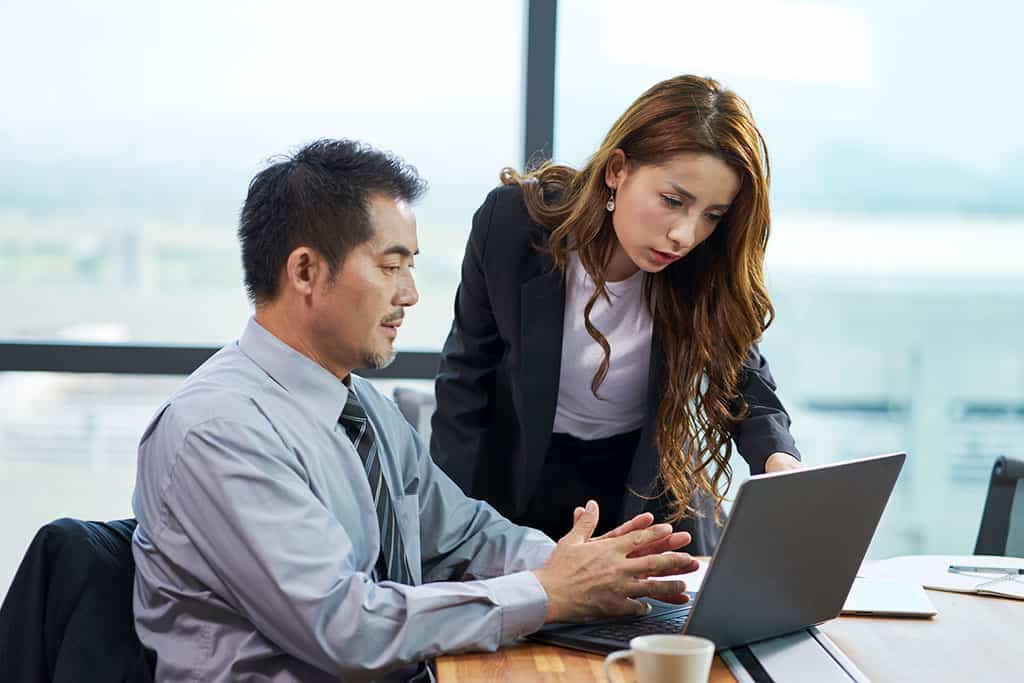 An Asian man and woman. The man is sitting at a desk in front of a computer. The woman is standing next to him while pointing at the computer screen.