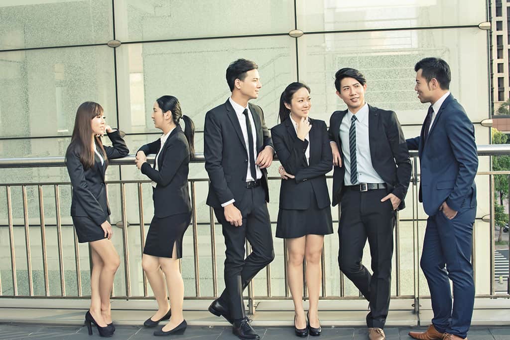A group of business people (4 men and 2 women) talking to each other. The two women are talking in one group, and the 4 men are talking in their own group.