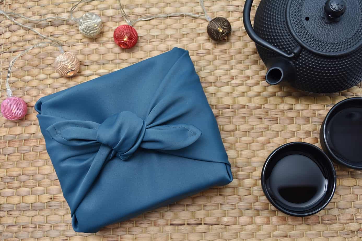 A box wrapped in a blue Furoshiki, or Japanese wrapping cloth on the left side, with an iron teapot and two cups to the right. There are also some Christmas looking ball decorations above the wrapped box.