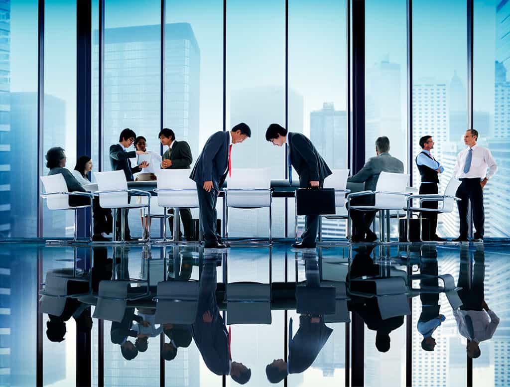 An office with glass windows that can see through to the outside. People are talking to each other, with two men in the center, facing each other and bowing.