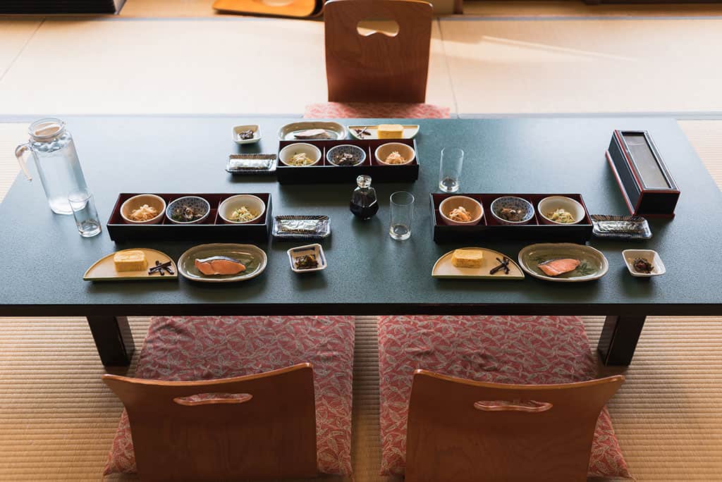 A low table in a tatami style Japanese room with chairs that sit directly on the ground. On the table are three meals made up a different dishes like salmon, egg, and assorted vegetables.