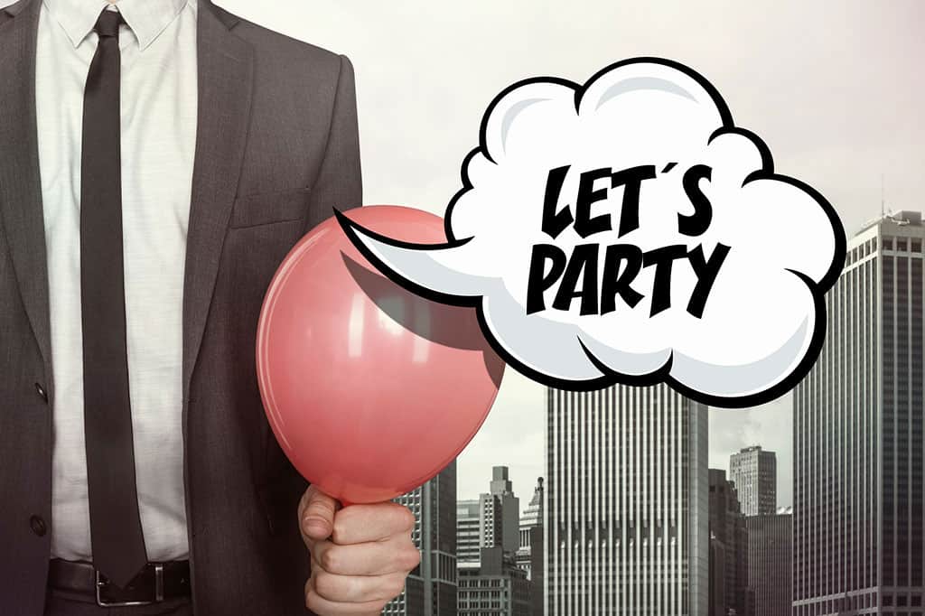 An image of a man (only his torso is visable) wearing a suit and tie holding a red balloon. There are buildings in the background and a comic speech bubble that has text that reads, "LET'S PARTY"