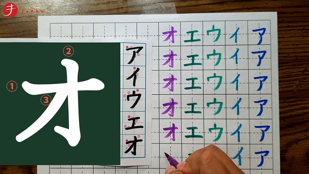 The katakana character "o" is shown on the left side of the image. On the right, a girl's hand can be seen writing the katakana characters "a, i, u, e, o" in different colors of ink with a pen on the right.