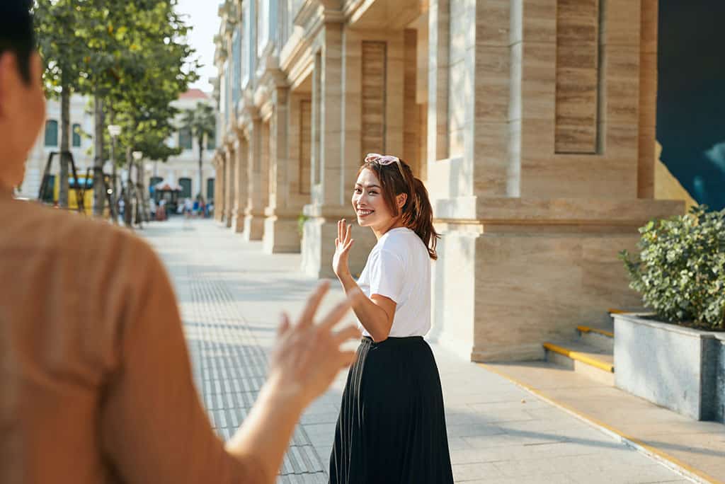 A young Asian girl walking away from a man. She is turning towards the man and waving goodbye and smiling.