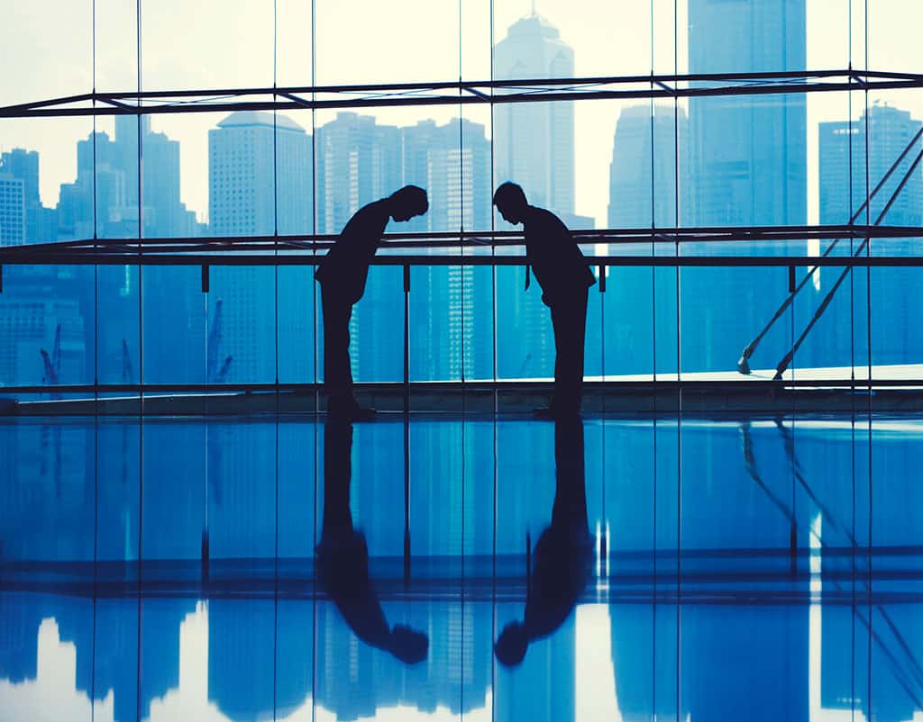 Silhouettes of two men can be seen bowing towards each other. They are in a building with glass windows, and in the background, other tall buildings can be seen.