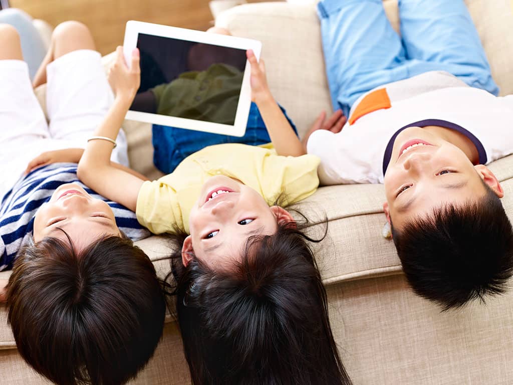 Three young children lying down upside down on a couch, so their heads are normally where your feet would be. The child in the middle is holding a smart device tablet.