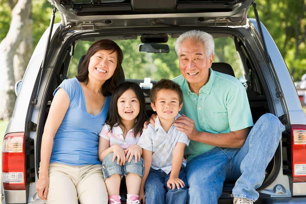 An elderly Asian woman and man with two young Asian children sitting in between them. The are sitting on the bummer of the back of a car with the trunk open.